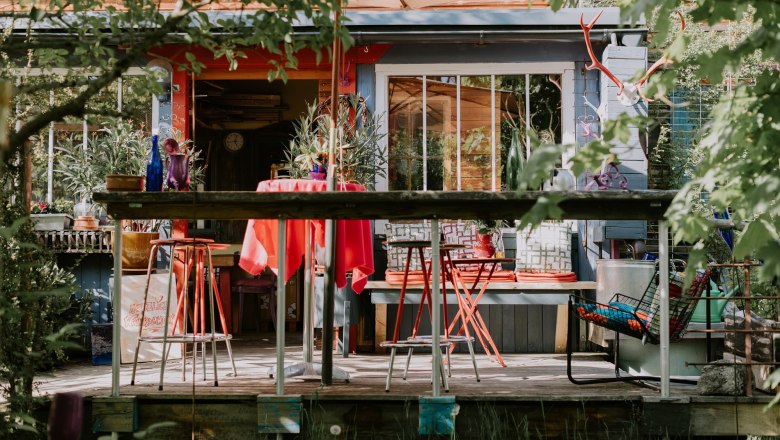 Bunte Terrasse vor der Location mit roten Stühlen und Tisch, umgeben von Pflanzen und Dekorationen.