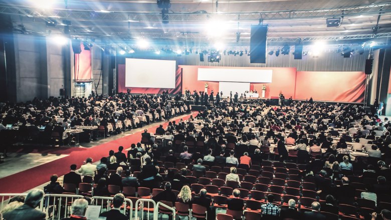 Großer Kongresssaal mit vielen Teilnehmern, Bühne und Leinwand.