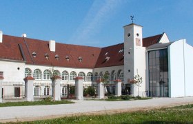 Schloss Katzelsdorf mit modernem Anbau und historischem Gebäude. Weiße Fassade mit gerundeten Fenstern, rotes Dach und großer Innenhof
