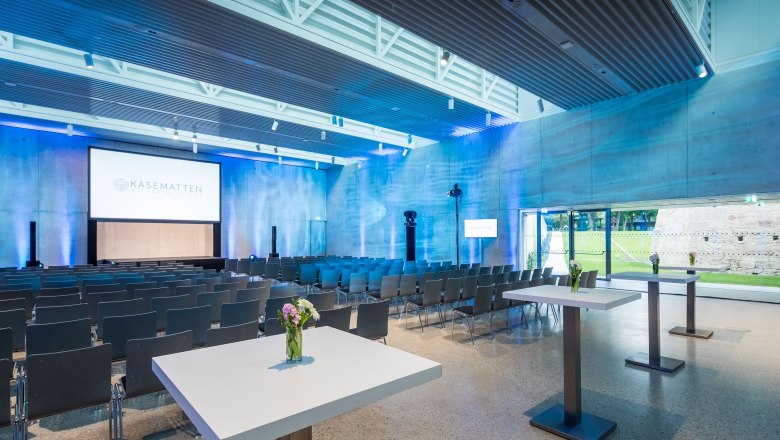 Moderner Konferenzraum mit St&uuml;hlen, Tischen und Leinwand, beleuchtet in Blau.