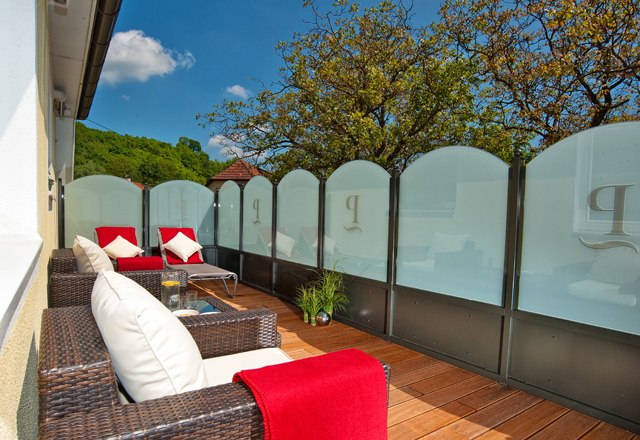 Eine Terrasse mit Rattanm&ouml;beln, roten Decken und Kissen, umgeben von Glasw&auml;nden und B&auml;umen im Hintergrund.