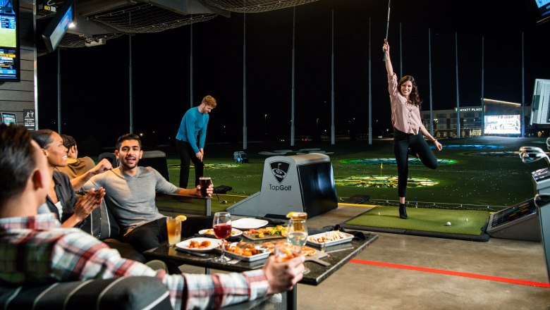 Menschen spielen Golf bei TopGolf, eine Frau jubelt, w&auml;hrend andere essen und trinken.