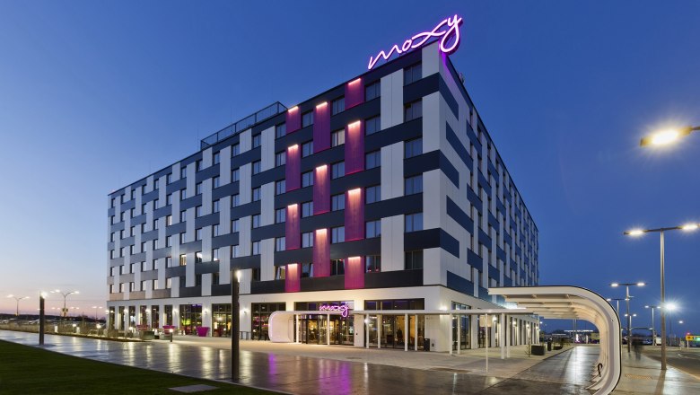 Au&szlig;enansicht des Moxy Vienna Airport Hotels bei D&auml;mmerung mit beleuchtetem Schriftzug.