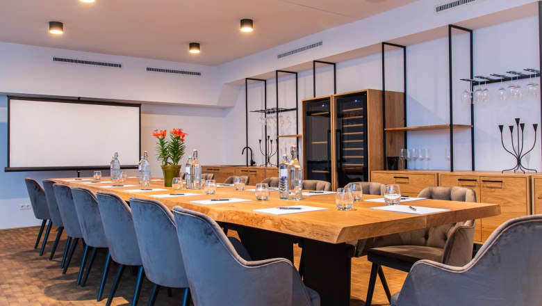 Moderner Konferenzraum mit großem Holztisch, grauen Stühlen und Präsentationsleinwand.