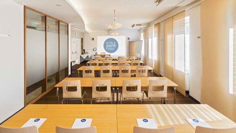 Ein Konferenzraum mit Tischen und St&uuml;hlen, einem Projektorbildschirm und Notizbl&ouml;cken mit dem Logo "at the park hotel".