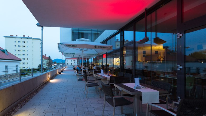 Terrasse eines Restaurants mit Tischen und St&uuml;hlen, beleuchtet von roten Lichtern, mit Blick auf Geb&auml;ude im Hintergrund.