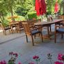 Ein gem&uuml;tlicher Gastgarten mit Holzm&ouml;beln und roten Sonnenschirmen, umgeben von gr&uuml;nen B&auml;umen und Blumen.