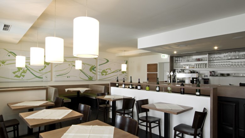 Innenansicht eines modernen Restaurants mit Holztischen, St&uuml;hlen und einer Bar mit Flaschen und Gl&auml;sern.