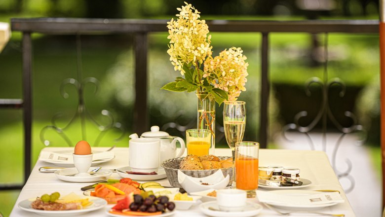 Frühstückstisch auf Terrasse mit Blumen, Saft, Brötchen und Eiern.