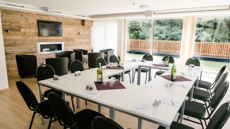 Konferenzraum mit U-f&ouml;rmiger Tischanordnung, St&uuml;hlen, Holzwand, Fernseher und gro&szlig;en Fenstern mit Blick ins Gr&uuml;ne.