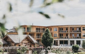 Ein modernes Holzgebäude mit Balkonen und ein traditionelles Holzhaus im Vordergrund, umgeben von Bäumen und Gartenanlagen.