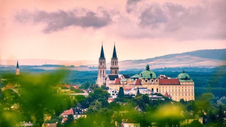 Panoramablick auf das Stift Klosterneuburg mit zwei Kirchtürmen und barockem Gebäude, umgeben von grüner Landschaft und unter bewölktem Himmel.