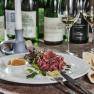 Ein Teller mit Schinken, Salat und Chutney, umgeben von Weinflaschen und Gl&auml;sern auf einem Tisch.