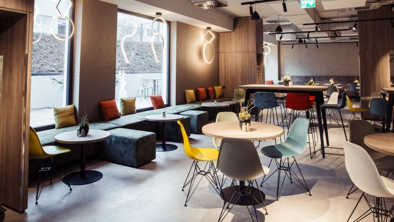 Modernes Hotel-Restaurant mit bunten Stühlen und Tischen, großen Fenstern und stilvollen Leuchten.