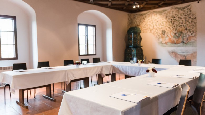 Tagungsraum mit U-f&ouml;rmiger Tischanordnung, wei&szlig;en Tischdecken und Schreibmaterialien. Im Hintergrund ein Wandgem&auml;lde und ein Kachelofen.