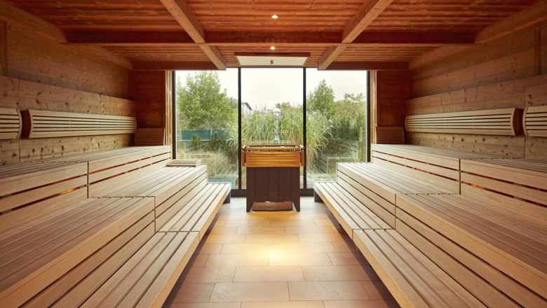 Innenansicht einer Sauna mit Holzb&auml;nken und einem Fenster mit Blick ins Gr&uuml;ne.
