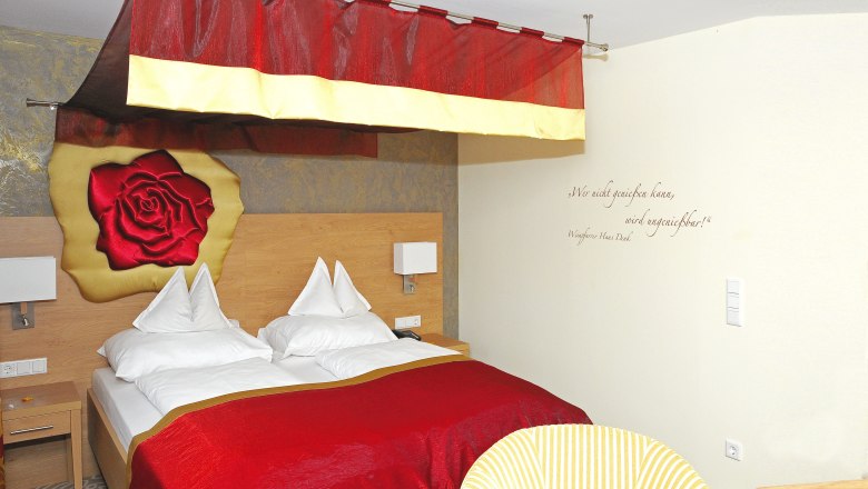 Gem&uuml;tliches Hotelzimmer mit rotem Baldachin und Rosenmotiv an der Wand.