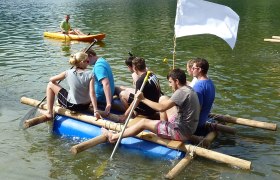 Gruppe von Menschen auf einem selbstgebauten Floß auf einem See, mit einer weißen Flagge und einem Kajakfahrer im Hintergrund.