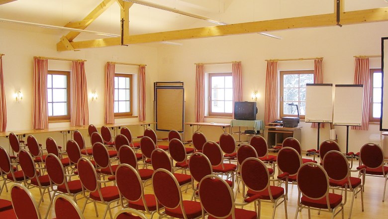 Ein Konferenzraum mit roten St&uuml;hlen, Holzbalken und Fenstern mit gestreiften Vorh&auml;ngen.
