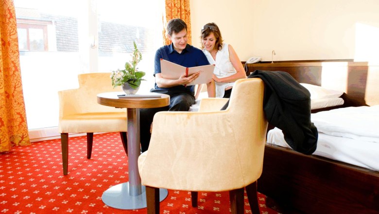 Ein Paar sitzt in einem Hotelzimmer auf einem Bett und schaut in ein Buch. Der Raum hat rote Teppiche und gelbe Vorh&auml;nge.