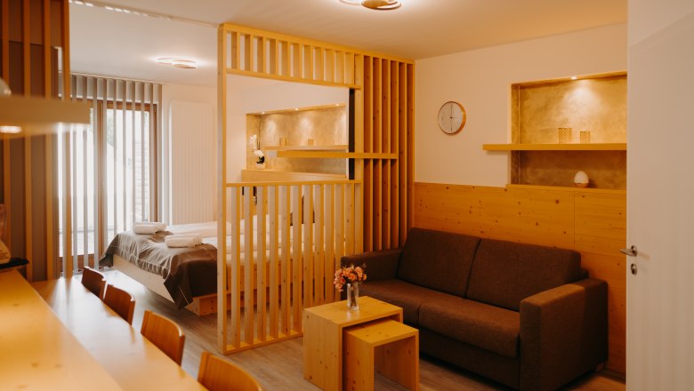 Modernes Hotelappartement mit Doppelbett, hellen Holzm&ouml;beln, ausziehbarer Couch und Raumtrenner aus Holz. Die Wand ist zum Teil mit Holz verkleidet und in der Wand sind Nischen, die in einem erdigen Gold gestrichen sind.