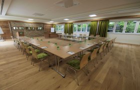 Seminarraum mit U-f&ouml;rmiger Tischanordnung, gr&uuml;nen St&uuml;hlen und gro&szlig;en Fenstern.