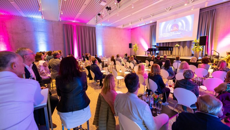 Menschen sitzen in einem modernen Veranstaltungsraum mit B&uuml;hne und Leinwand.