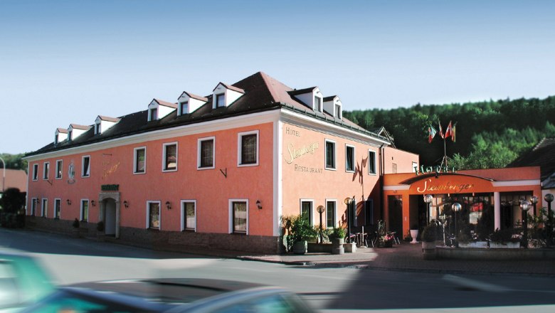 Ein rosafarbenes Hotelgeb&auml;ude mit mehreren Fenstern und einem Eingang mit Vordach. Im Vordergrund f&auml;hrt ein Auto vorbei.