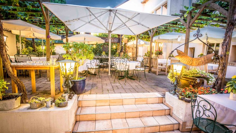 Gem&uuml;tlicher Gastgarten mit Tischen, St&uuml;hlen und Sonnenschirmen, umgeben von Pflanzen und Lichtern.