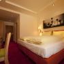 Ein gem&uuml;tliches Hotelzimmer mit Doppelbett, Schreibtisch und Wandbild. Warme Beleuchtung und rote Teppiche schaffen eine einladende Atmosph&auml;re.