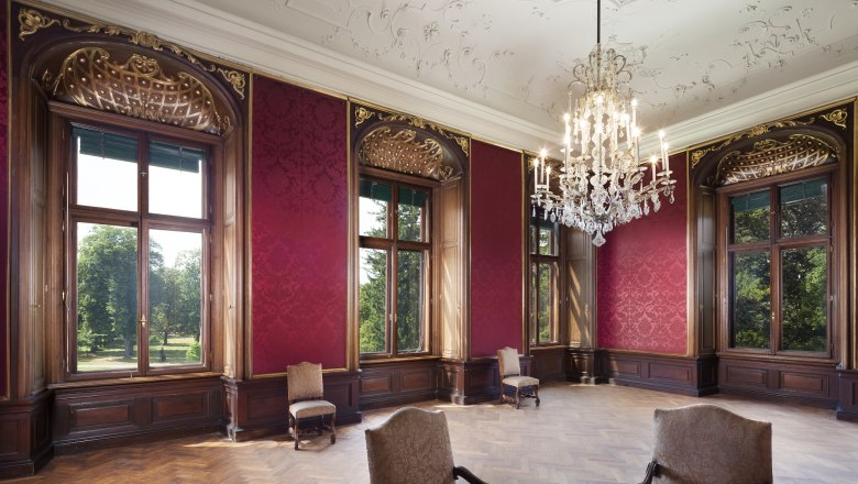 Eleganter Raum im Schloss Eckartsau mit roten Wänden, großen Fenstern und einem Kronleuchter.