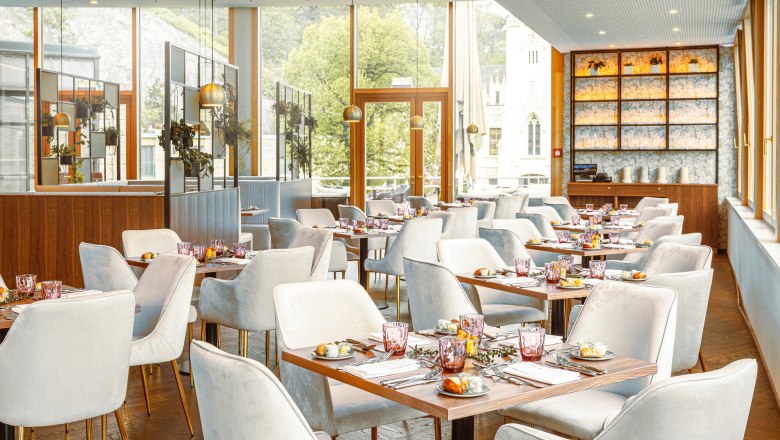 Elegantes Restaurant mit gedeckten Tischen und weißen Stühlen, große Fenster mit Blick ins Grüne.