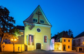 Gelbes Geb&auml;ude mit Kreuzsymbol, beleuchtet bei Nacht.