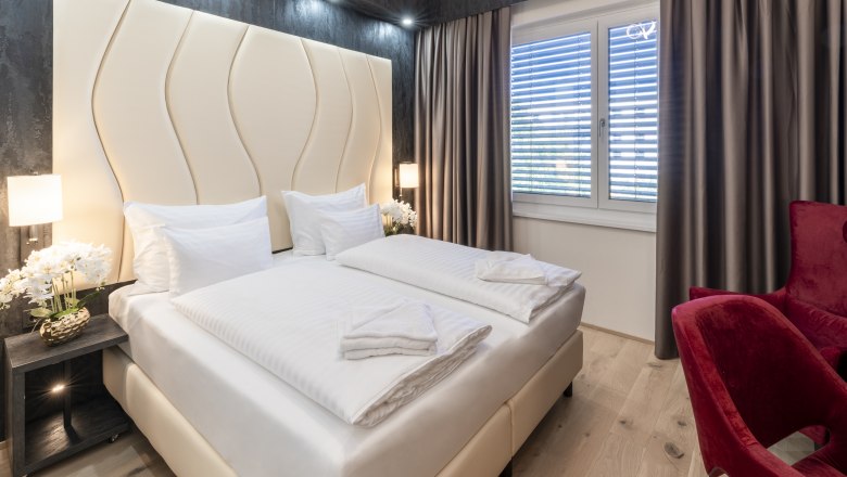 Modernes Hotelzimmer mit Doppelbett, roten Sesseln und gro&szlig;en Fenstern mit Vorh&auml;ngen.