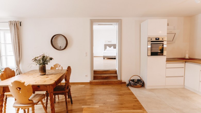 Helle Küche mit Holztisch, Stühlen und modernen weißen Schränken. Durchgang zu einem Schlafzimmer im Hintergrund.