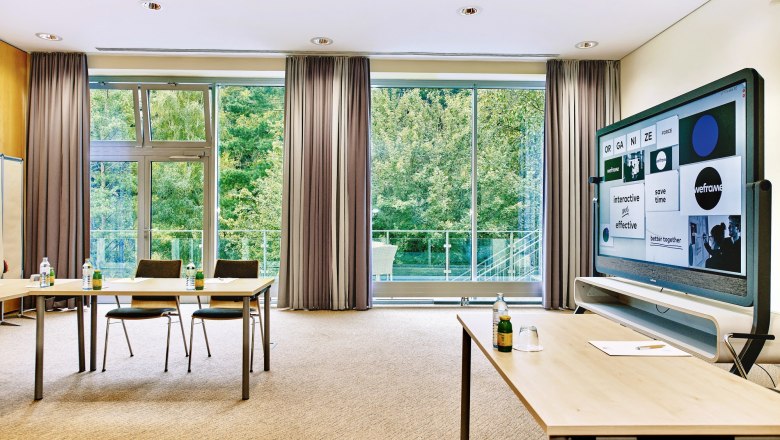 Moderner Seminarraum mit großen Fenstern, Tischen, Stühlen und einem interaktiven Bildschirm.