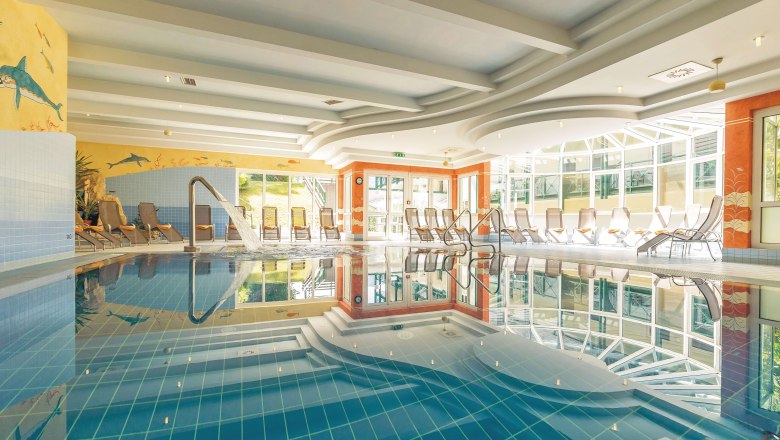 Ein helles Hallenbad mit Liegestühlen und einem Wasserstrahl im Pool.