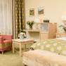 Gem&uuml;tliches Hotelzimmer mit gr&uuml;nem Teppich, rotem Sessel, Tisch mit Sektk&uuml;hler, Blumen und Bett mit gr&uuml;nem &Uuml;berwurf.