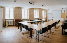 Ein heller Seminarraum mit U-f&ouml;rmiger Tischanordnung, St&uuml;hlen, Projektionsleinwand und Flipchart.