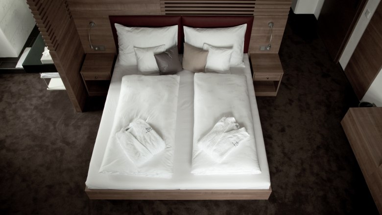 Ein modernes Hotelzimmer mit einem Doppelbett, wei&szlig;en Bettdecken und Badem&auml;nteln, umgeben von Holzm&ouml;beln.