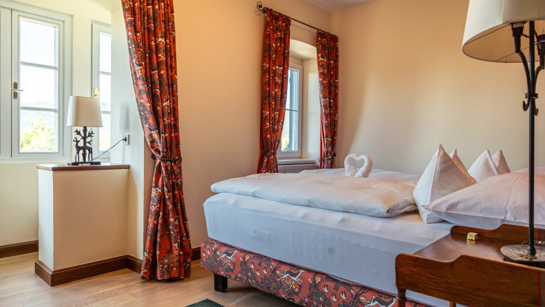 Gem&uuml;tliches Hotelzimmer mit Doppelbett, roten Vorh&auml;ngen und dekorativem Handtuch in Herzform.