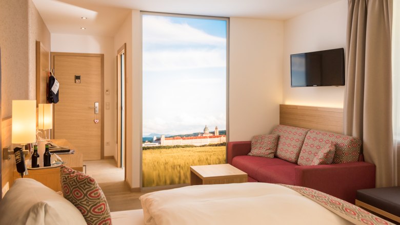 Modernes Hotelzimmer mit Doppelbett, rotem Sofa, Flachbildfernseher und großem Fenster mit Landschaftsausblick.