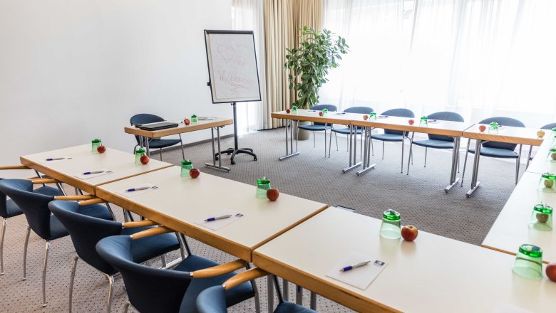 Konferenzraum mit Tischen in U-Form, Stühlen, Flipchart und Schreibmaterialien.