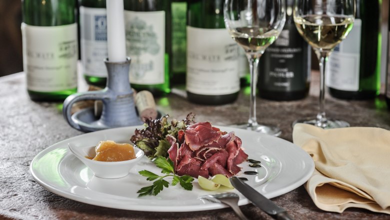 Ein Teller mit Schinken, Salat und Chutney, umgeben von Weinflaschen und Gläsern auf einem Tisch.