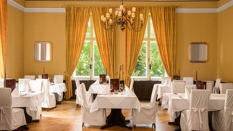 Elegantes Hotelrestaurant mit gedeckten Tischen, weißen Stuhlhussen und großen Fenstern mit gelben Vorhängen.