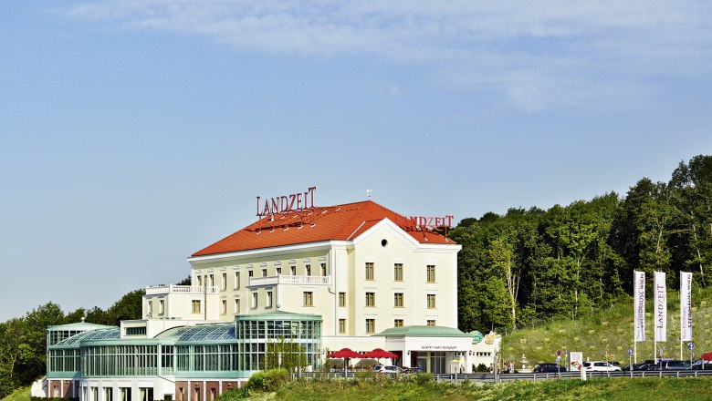 Landzeit Steinhäusl Gebäude mit rotem Dach und Glasfassade, umgeben von Bäumen und blauen Himmel.