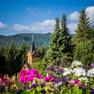 Kirchturm inmitten von B&auml;umen und Blumen vor einer Berglandschaft.