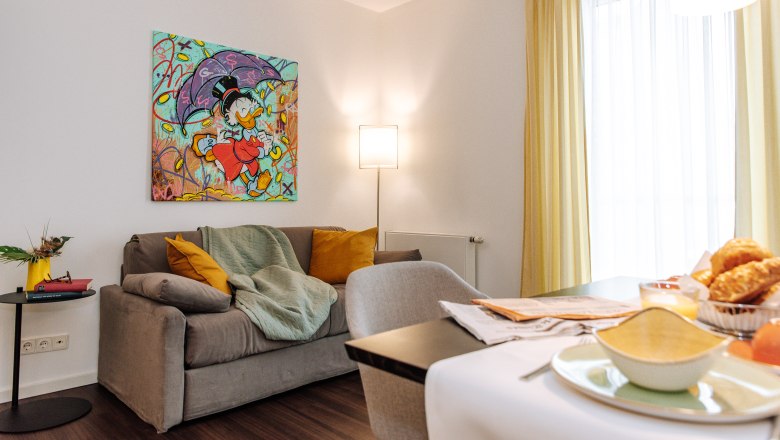 Wohnzimmer mit grauem Sofa, bunter Comic-Kunst an der Wand, gelben Vorhängen und Frühstückstisch.