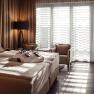 Hotelzimmer mit Doppelbett, Badem&auml;nteln, zwei Sesseln und einem Tisch vor einem gro&szlig;en Fenster mit Vorh&auml;ngen.