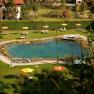 Ein nat&uuml;rlicher Schwimmteich mit Liegest&uuml;hlen und bunten Sonnenschirmen auf einer gr&uuml;nen Wiese.